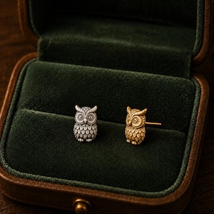 May include: A pair of owl-shaped stud earrings, one in a silver-toned finish and one in a gold-toned finish, featuring detailed feather textures and large circular eyes. The earrings are displayed side by side on a dark green velvet surface.