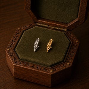 May include: Two small metal pins shaped like wheat stalks, one in a silver tone and one in a gold tone, resting inside an octagonal wooden box with a dark green velvet lining. The pins feature detailed textures resembling individual grains.
