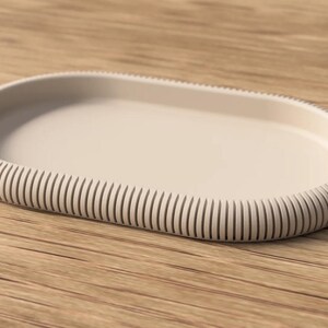 Könnte beinhalten: Ein ovales Tablett in einem einfarbigen Hellbeige. Das Tablett verfügt über eine glatte, flache Innenfläche und einen erhöhten, dicken Außenrand, der mit einer Reihe vertikaler, paralleler Rillen rund um den gesamten Umfang verziert ist.