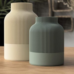 Könnte beinhalten: Zwei zylindrische Vasen mit vertikal gerippten Texturen im oberen Bereich und glatten Unterteilen. Die größere Vase ist cremefarben, die kleinere ist salbeigrün. Beide haben einen schmalen Hals und eine breite, flache Öffnung oben.