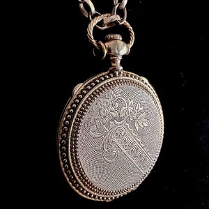 May include: A silver-toned circular pendant on a thick chain, featuring an intricate engraved floral pattern and a fine mesh-like texture on the surface. The pendant has a beaded border and a decorative top loop connecting it to the metal chain.