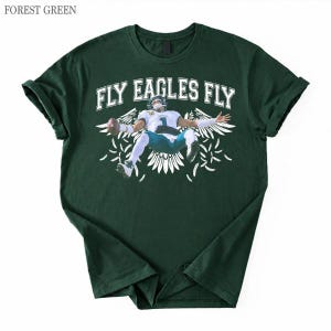 May include: A forest green short-sleeve t-shirt featuring a graphic of a football player in a white jersey and teal trousers holding a football. The text FLY EAGLES FLY is printed in white above the player, surrounded by white wing illustrations.