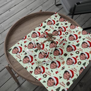 May include: A light green wrapping paper featuring a repeating pattern of a smiling person wearing a Santa hat, small Christmas trees, red stockings, wreaths, and the text Merry Christmas, tied with a brown ribbon and a bow.