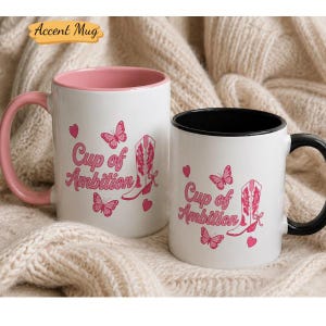 May include: Two white ceramic mugs featuring pink graphics of a cowboy boot, butterflies, and hearts. The text "Cup of Ambition" is printed in pink. One mug has a pink handle and interior, while the other has a black handle and interior.