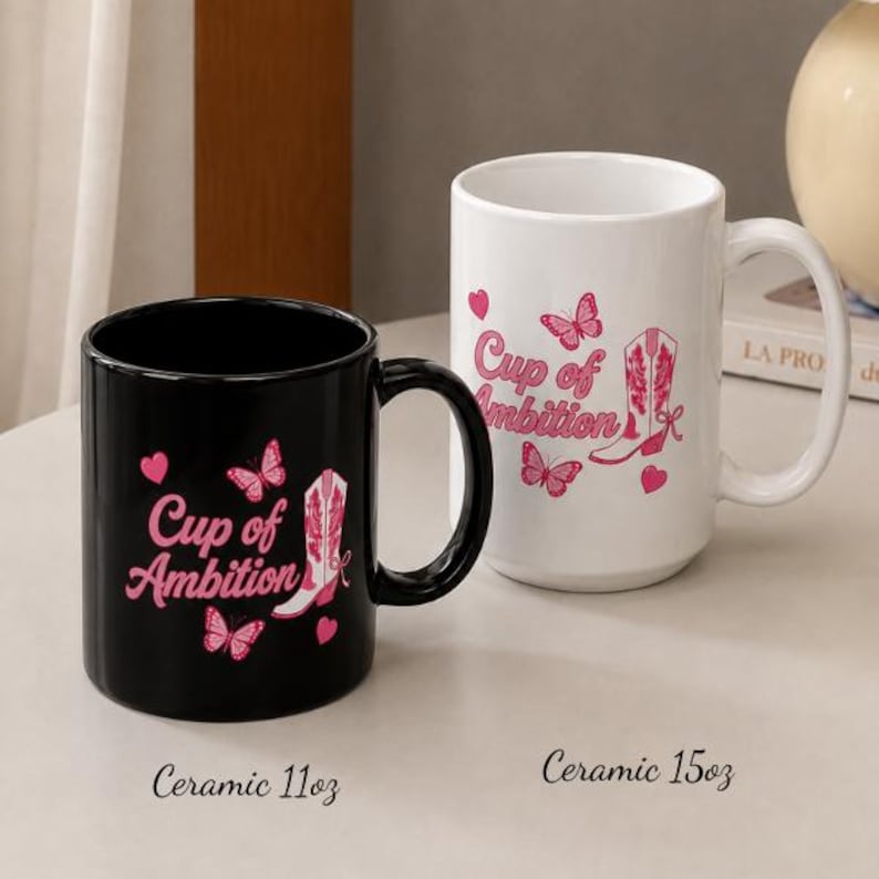 May include: A black ceramic 11oz mug and a white ceramic 15oz mug, both featuring the pink text Cup of Ambition, pink butterfly illustrations, and a pink cowboy boot graphic. The mugs are displayed side by side on a flat surface.