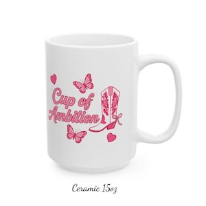 May include: A white ceramic mug with a 425 ml capacity, featuring a pink graphic design that includes the text "Cup of Ambition," a western-style boot, two butterflies, and two small hearts printed on the front surface of the mug.