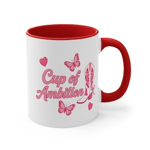 May include: A white ceramic mug with a red interior and handle. The exterior features the pink text "Cup of Ambition" alongside illustrations of a pink cowboy boot, two pink butterflies, and two small pink hearts arranged on the white surface.
