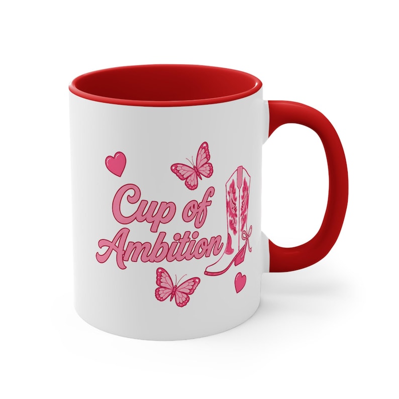 May include: A white ceramic mug with a red interior and handle. The exterior features the pink text "Cup of Ambition" alongside illustrations of a pink cowboy boot, two pink butterflies, and two small pink hearts arranged on the white surface.