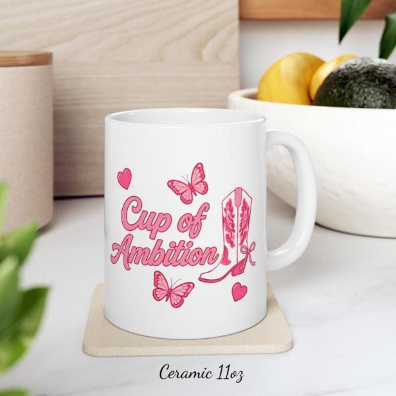 May include: A white ceramic mug featuring pink graphics of two butterflies, two small hearts, and a western-style boot. The text "Cup of Ambition" is printed in a pink cursive font on the front. The mug has a curved handle and sits on a square coaster.
