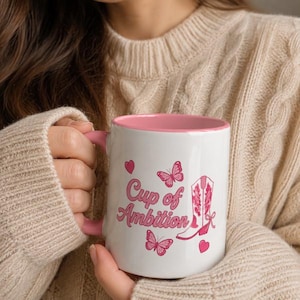 May include: A white ceramic mug with a pink interior and handle. The exterior features the pink text "Cup of Ambition" alongside pink butterfly illustrations and a pink western-style cowboy boot graphic with decorative floral patterns.