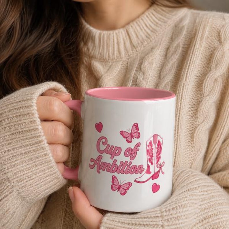 May include: A white ceramic mug with a pink interior and handle. The exterior features the pink text "Cup of Ambition" alongside pink butterfly illustrations and a pink western-style cowboy boot graphic with decorative floral patterns.