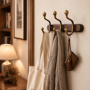 May include: A wall-mounted coat rack featuring a dark brown wooden base with three brass-toned metal hooks. Each hook has a curved design with a rounded finial at the top and bottom. A beige canvas tote bag, a grey knit scarf, and a brown leather key fob hang.