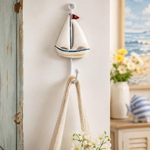 May include: A white wall-mounted hook featuring a decorative ceramic sailboat ornament. The sailboat has white sails, a small red flag at the top, and yellow and blue painted accents along the hull. A beige fabric tote bag hangs from the hook.