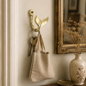 May include: A gold-coloured metal wall hook shaped like a whale tail. The hook is mounted to the wall and holds a silver metal keyring with several keys attached, along with a small, beige, rectangular fabric bag with two short handles.