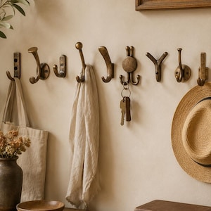 Peut inclure: Une collection de huit patères murales en laiton de style ancien, fixées en rangée horizontale. Les crochets présentent divers designs incurvés et à plusieurs branches, dont l'un porte un trousseau de clés. La finition est un ton doré métallique.