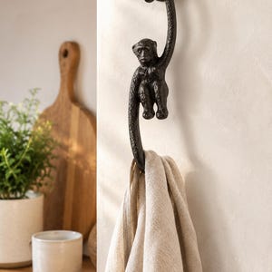 Op de afbeelding: Een donkerbruine, getextureerde metalen wandhaak in de vorm van een aap die aan zijn staart hangt. De haak is bevestigd aan een lichtgekleurde muur en houdt een beige linnen handdoek vast die over de onderste bocht van de staart hangt.