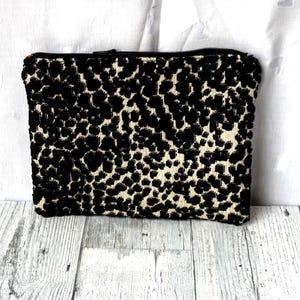 May include: A rectangular clutch with a black and cream leopard print pattern. The clutch has a black zipper closure and is made of a textured fabric. The bag is a neutral accessory.