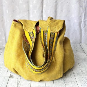 May include: A mustard yellow tote bag with a decorative strap. The bag has a flap closure and a patterned strap with yellow, blue, and red accents. The bag is made of a textured fabric.
