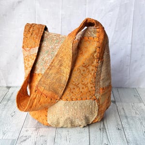 Peut inclure: Un sac fourre-tout orange avec un motif patchwork. Le sac présente une variété de tissus, y compris des matériaux à motifs et texturés. Les bretelles sont larges et en tissu orange uni. Le sac est posé sur une surface en bois clair.