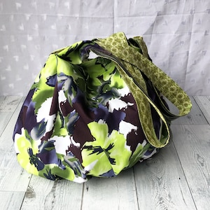 May include: A large, green and purple floral print tote bag with two green and brown straps. The bag is made of fabric and has a casual, relaxed style.