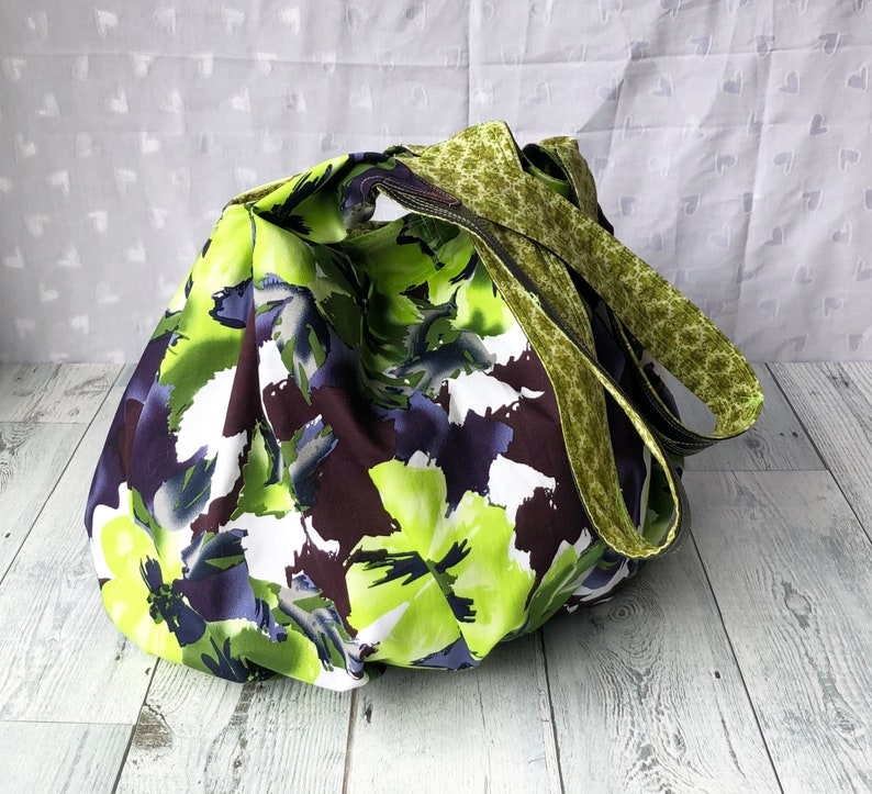 May include: A large, green and purple floral print tote bag with two green and brown straps. The bag is made of fabric and has a casual, relaxed style.