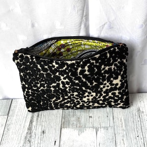May include: A black and white patterned clutch bag with a zipper closure. The exterior features a textured, spotted design, while the interior lining showcases a colorful, abstract pattern. The bag is open, revealing the vibrant interior.