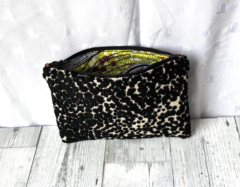 May include: A black and white patterned clutch bag with a zipper closure. The exterior features a textured, spotted design, while the interior lining showcases a colorful, abstract pattern. The bag is open, revealing the vibrant interior.