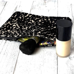 May include: A black and white leopard print cosmetic bag with a black zipper, a black makeup brush, and a bottle of foundation. The foundation bottle has a black cap and a beige base. The bag is open, revealing a colorful interior.