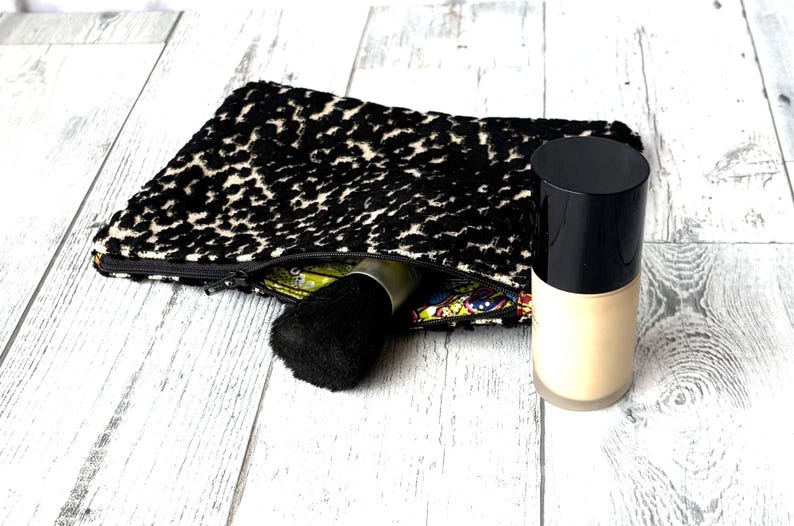 May include: A black and white leopard print cosmetic bag with a black zipper, a black makeup brush, and a bottle of foundation. The foundation bottle has a black cap and a beige base. The bag is open, revealing a colorful interior.