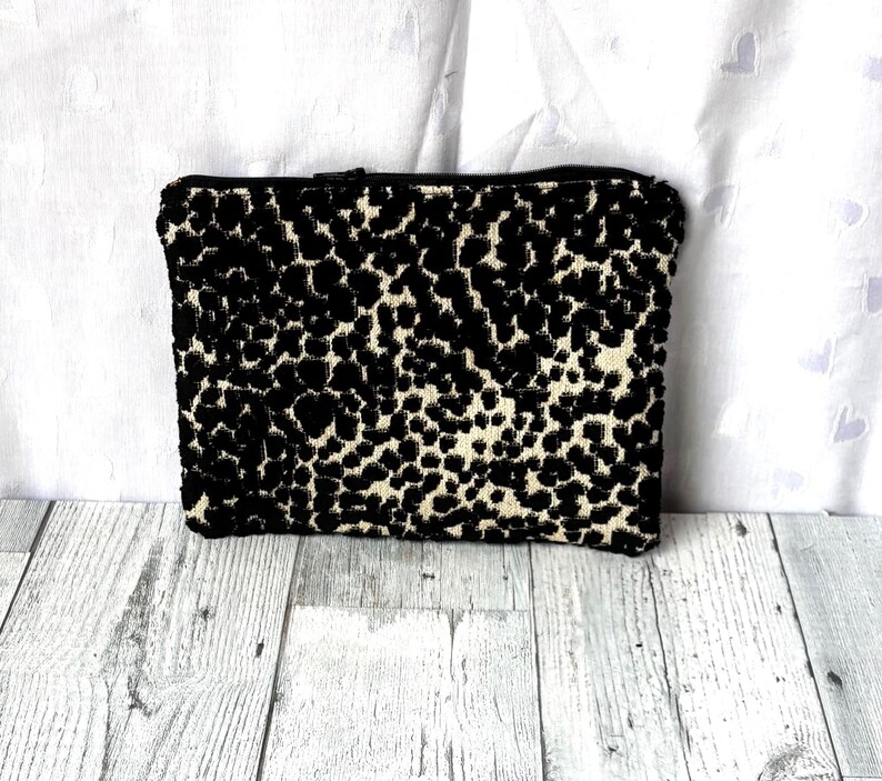May include: A rectangular clutch with a black and cream leopard print pattern. The clutch has a black zipper closure and is made of a textured fabric. The bag is a neutral accessory.