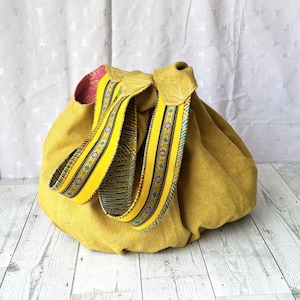May include: Mustard yellow drawstring bag with decorative yellow straps. The straps feature a colorful pattern with blue, red, and green accents. The bag's interior is lined with a pink fabric.