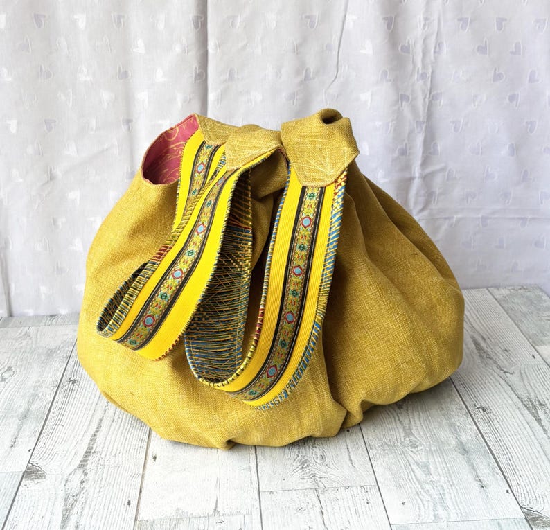 May include: Mustard yellow drawstring bag with decorative yellow straps. The straps feature a colorful pattern with blue, red, and green accents. The bag's interior is lined with a pink fabric.