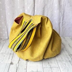 May include: Mustard yellow fabric bag with a decorative yellow strap featuring a colorful geometric pattern. The bag has a rounded shape and a contrasting burgundy interior. The strap is folded over the top of the bag.