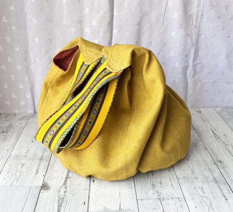 May include: Mustard yellow fabric bag with a decorative yellow strap featuring a colorful geometric pattern. The bag has a rounded shape and a contrasting burgundy interior. The strap is folded over the top of the bag.