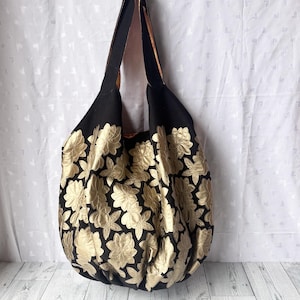 Op de afbeelding: Een grote zwarte tote bag met gouden bloemenborduurwerk. De tas heeft een enkele band en is gemaakt van een zachte, gestructureerde stof.