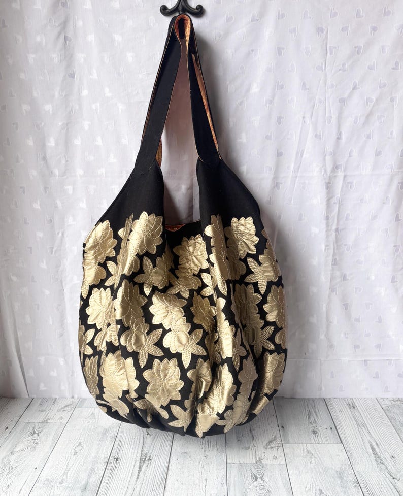 Op de afbeelding: Een grote zwarte tote bag met gouden bloemenborduurwerk. De tas heeft een enkele band en is gemaakt van een zachte, gestructureerde stof.
