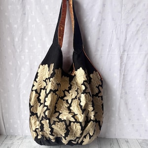 Op de afbeelding: Een grote zwarte tote bag met gouden bloemenborduurwerk. De tas heeft een bruin en zwarte band.