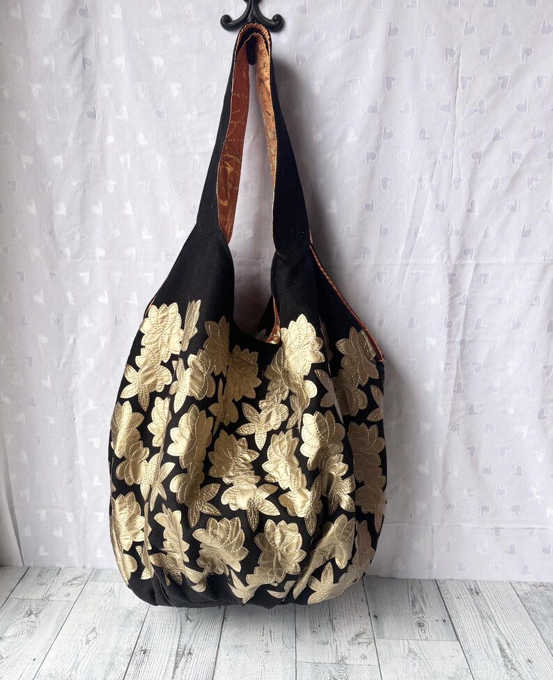 Op de afbeelding: Een grote zwarte tote bag met gouden bloemenborduurwerk. De tas heeft een bruin en zwarte band.