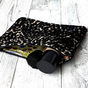 May include: A black and cream leopard print cosmetic bag with a zipper, partially open to reveal makeup brushes and containers. The bag's interior features a colorful patterned fabric. The bag is on a light wood surface.