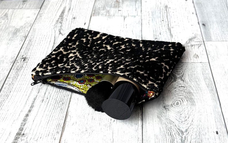 May include: A black and cream leopard print cosmetic bag with a zipper, partially open to reveal makeup brushes and containers. The bag's interior features a colorful patterned fabric. The bag is on a light wood surface.