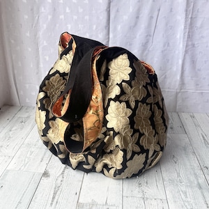Op de afbeelding: Een zwarte tote bag met gouden bloemenborduurwerk. De tas heeft een zwarte band en een bruine voering.