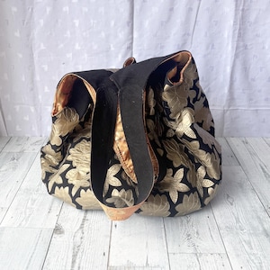 Op de afbeelding: Een zwarte en gouden tote bag met een bloemenpatroon. De tas heeft twee zwarte hengsels en een gouden voering.