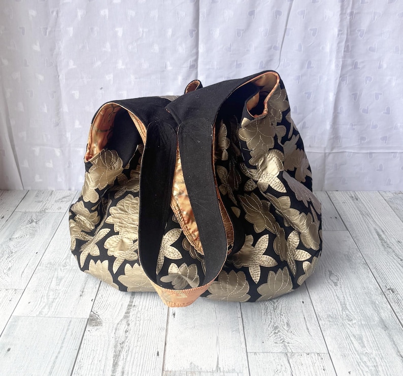 Op de afbeelding: Een zwarte en gouden tote bag met een bloemenpatroon. De tas heeft twee zwarte hengsels en een gouden voering.