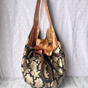 Op de afbeelding: Een grote zwarte en gouden tote bag met een bloemenpatroon. De tas heeft een lange band en is gemaakt van een zachte, gestructureerde stof. De binnenkant van de tas is gevoerd met een perzikkleurige stof.