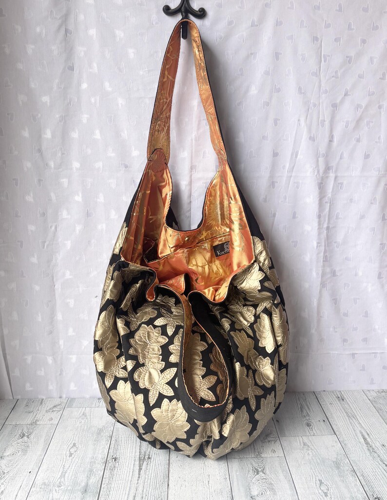 Op de afbeelding: Een grote zwarte en gouden tote bag met een bloemenpatroon. De tas heeft een lange band en is gemaakt van een zachte, gestructureerde stof. De binnenkant van de tas is gevoerd met een perzikkleurige stof.