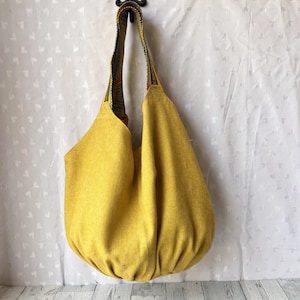 May include: A large, mustard-yellow tote bag with a rounded bottom and a patterned strap. The bag is made of a textured fabric and hangs from a decorative hook. The strap features a colorful, woven design.