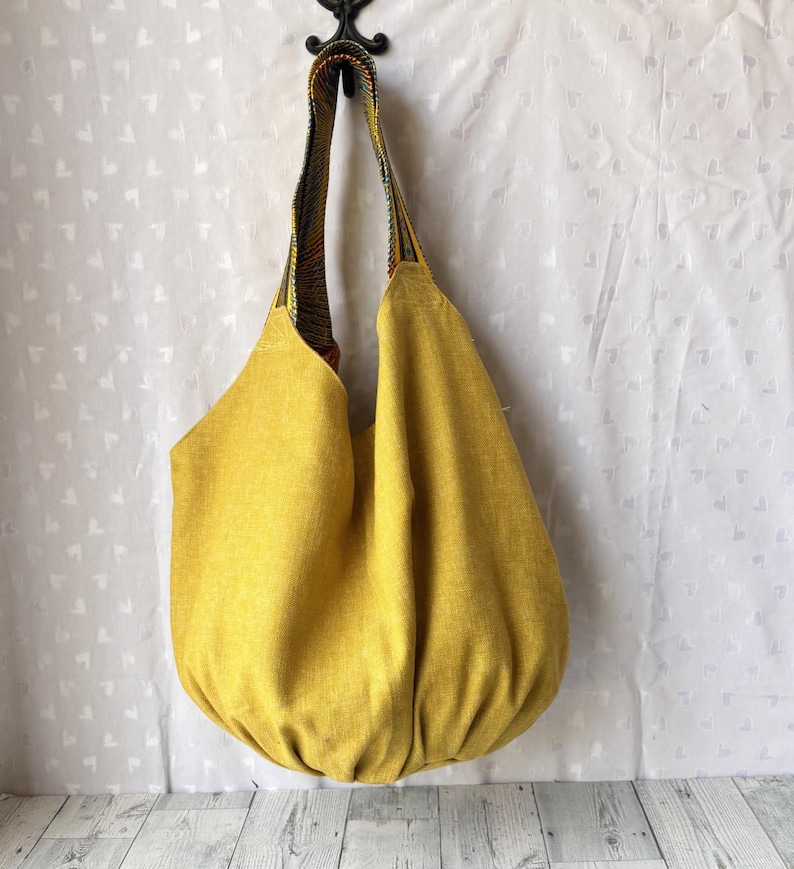 May include: A large, mustard-yellow tote bag with a rounded bottom and a patterned strap. The bag is made of a textured fabric and hangs from a decorative hook. The strap features a colorful, woven design.