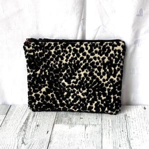 May include: A rectangular black and cream-colored clutch bag with a leopard print pattern. The bag has a zipper closure and is made of a textured fabric. The bag is on a white and gray wooden surface.