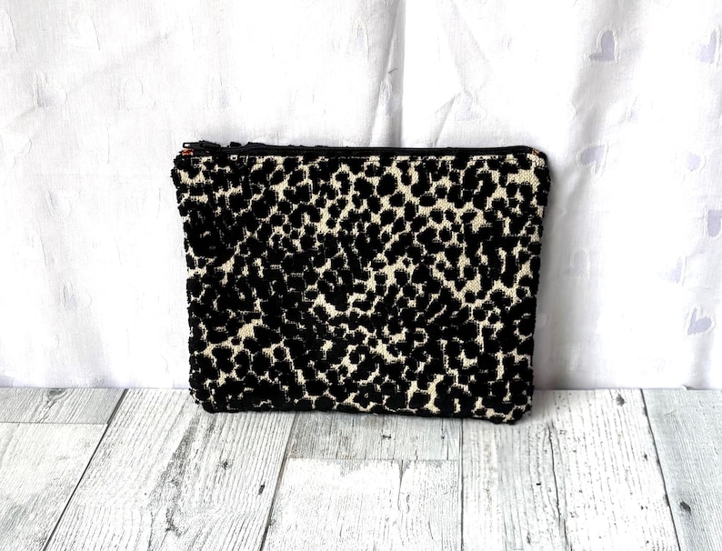 May include: A rectangular black and cream-colored clutch bag with a leopard print pattern. The bag has a zipper closure and is made of a textured fabric. The bag is on a white and gray wooden surface.