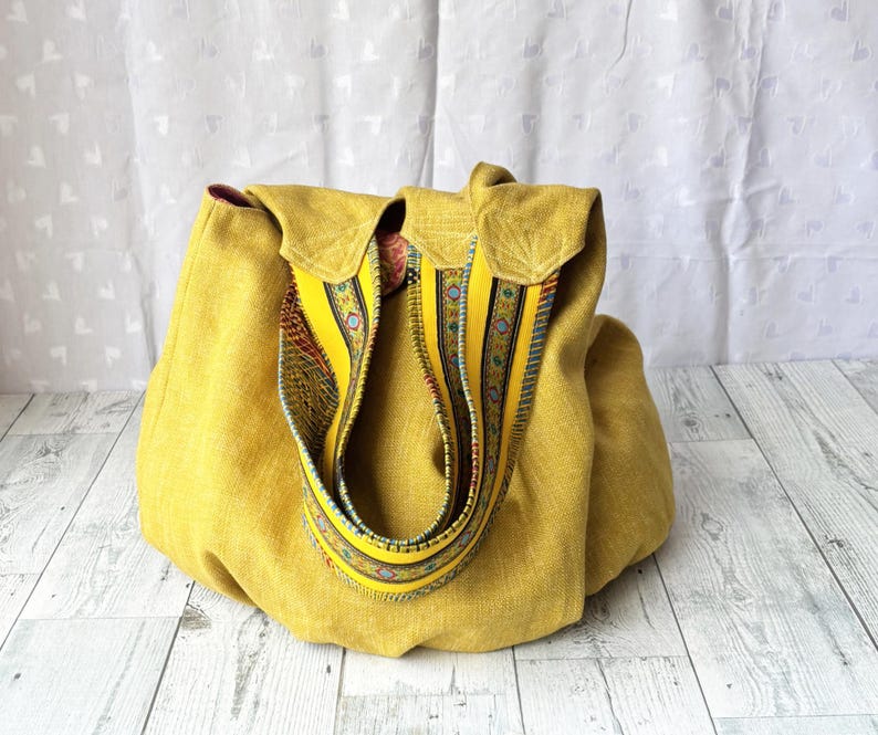 May include: A mustard yellow tote bag with a decorative strap. The bag has a flap closure and a patterned strap with yellow, blue, and red accents. The bag is made of a textured fabric.